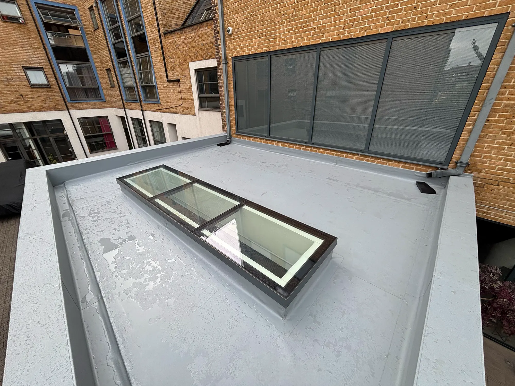 Flat roof with light grey waterproofing, large four-panel skylight, and surrounding brick buildings