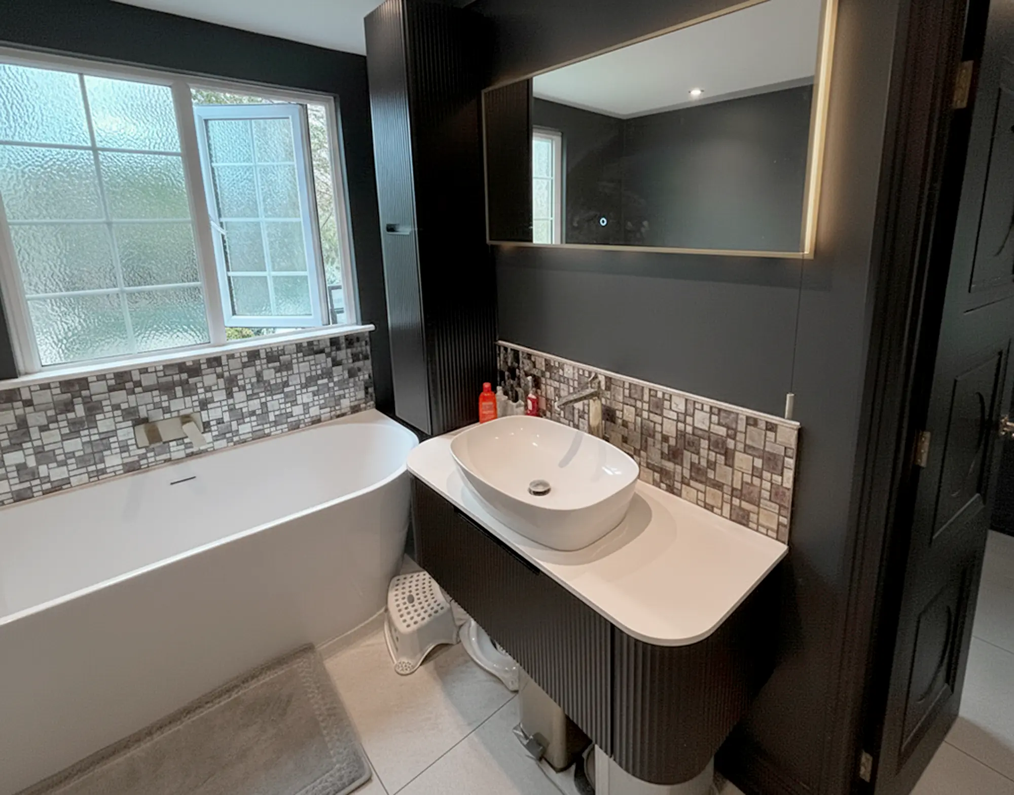 Modern bathroom with freestanding bath, dark walls, fluted vanity, and backlit mirror